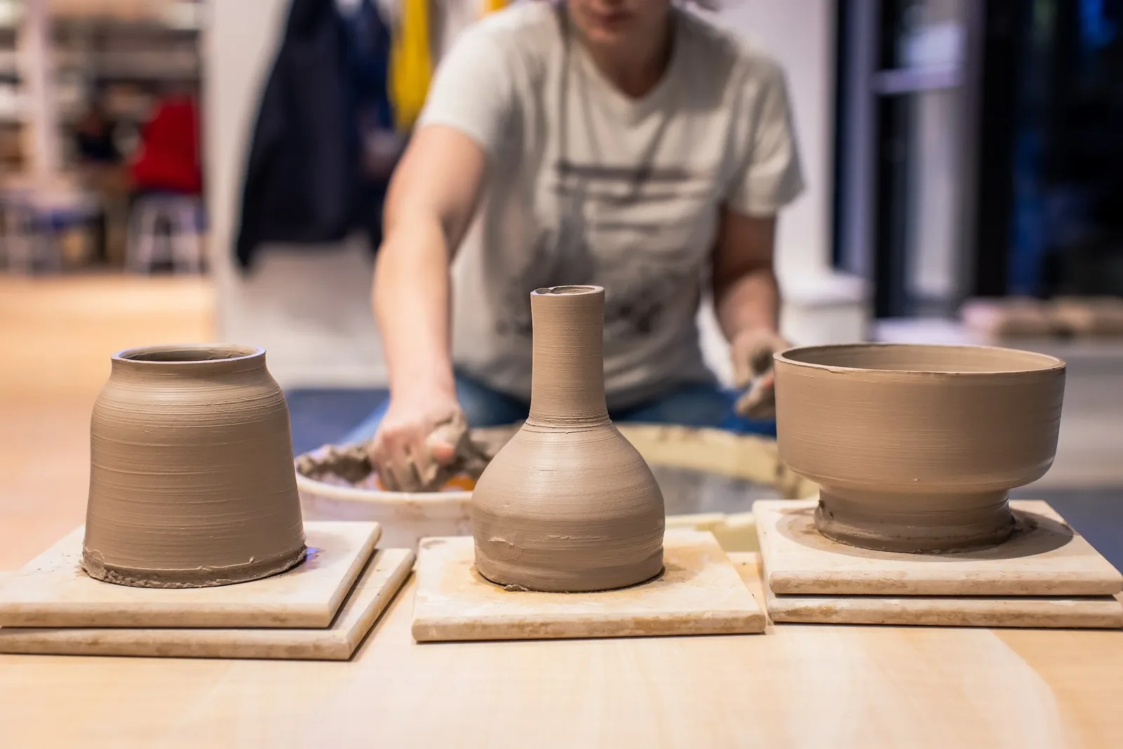 Three unfired pots on ware boards (a jar, a bottle vase and a footed bowl), with a potter at the wheel behind them