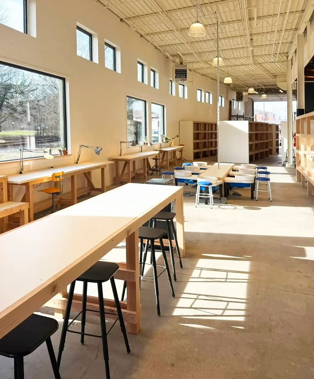 Sunlit ViaClay Member Studio: worktables along the windows, tall tables with stools and a row of pottery wheels