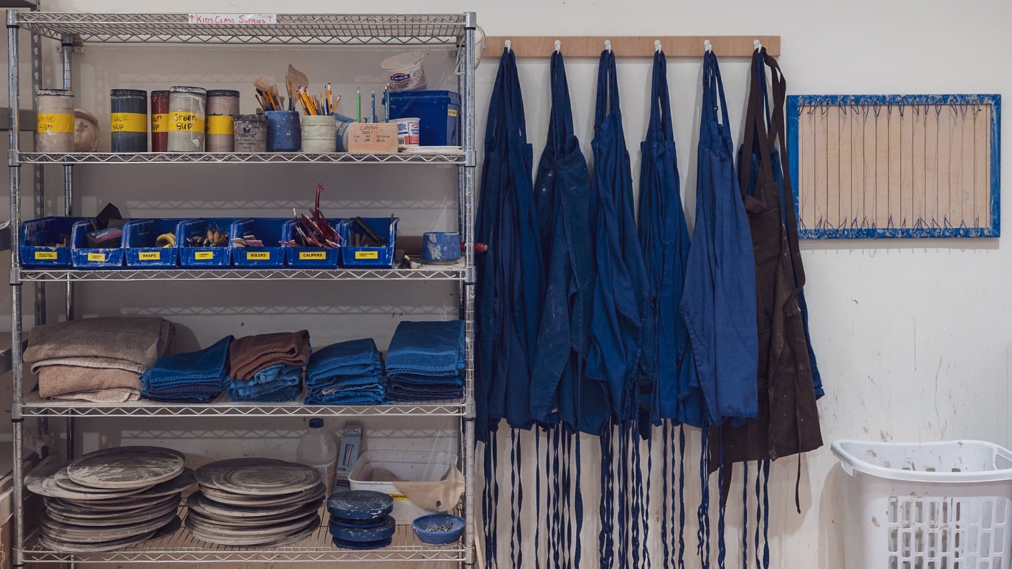 Blue studio aprons on hooks beside a shelf of clay tools, towels and wheel bats at ViaClay
